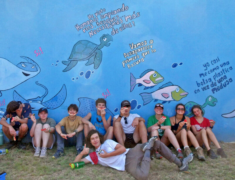 A group of people are sitting in front of a mural depicting marine life. The mural is painted in blue and features images of turtles, fish, and stingrays. There are also some Spanish phrases written on the mural, seemingly about protecting the ocean. The people are smiling and appear to be enjoying themselves. They are likely involved in some kind of environmental or conservation effort.